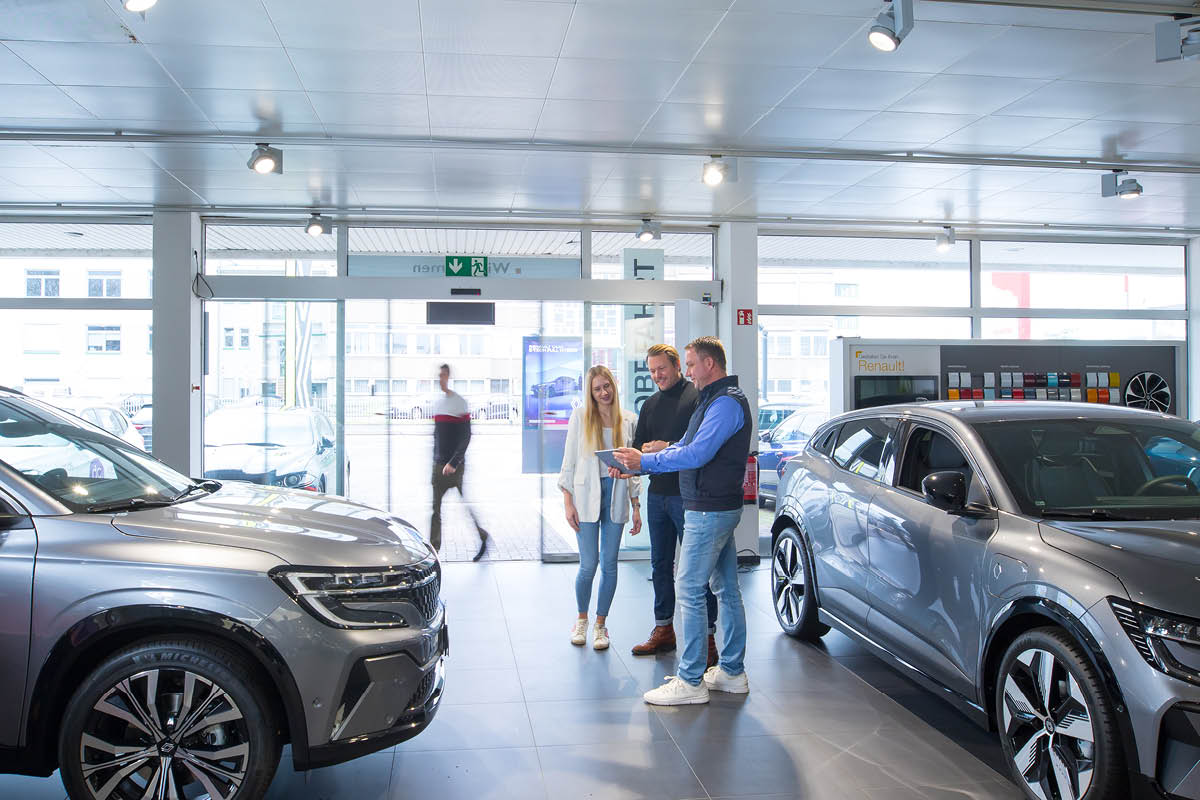 Innenraum eines modernen Autohauses. Drei Personen befinden sich in der Mitte und unterhalten sich, während der Mann im blauen Hemd auf ein Tablet zeigt.
