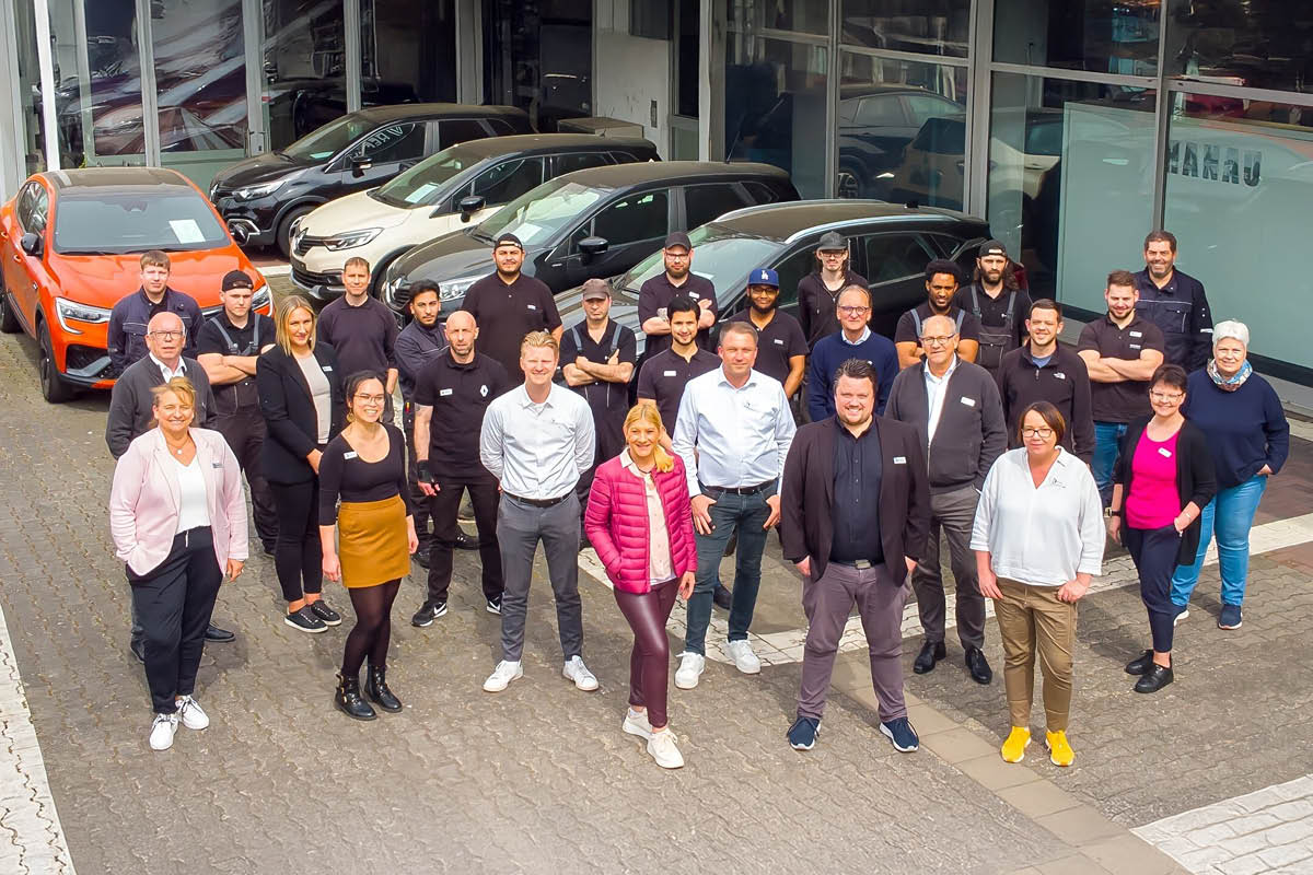 Eine Gruppe von Mitarbeiter steht vor einem Autohaus und posiert für ein Gruppenfoto. Im Hintergrund sind mehrere geparkte Autos zu sehen.