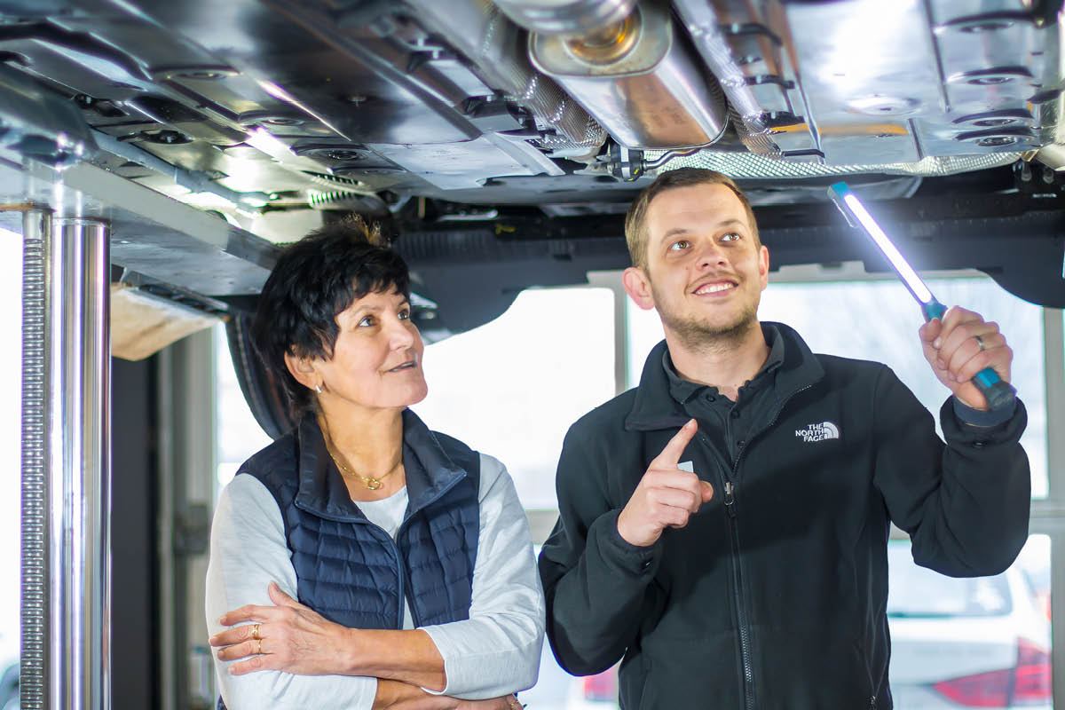 Zwei Personen stehen unter einem aufgebockten Auto in einer Werkstatt. Eine Person hält eine Lampe und zeigt auf einen Bereich unter dem Fahrzeug, während die andere aufmerksam zuhört.