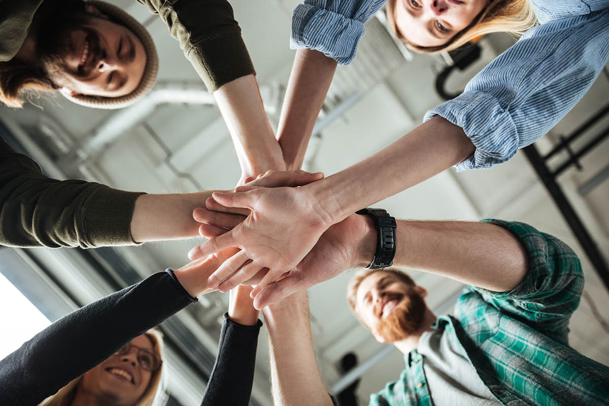 Eine Gruppe von Menschen, die ihre Hände in der Mitte übereinanderlegen. Die Perspektive ist von unten aufgenommen.